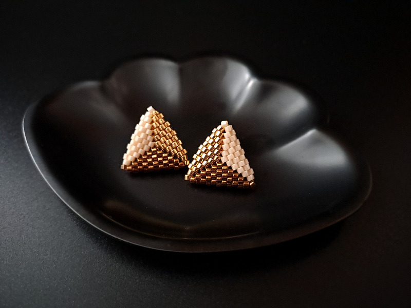 Beżowe trójkątne kolczyki z koralików w złotych i brązowych tonach, idealne na eleganckie wyjścia. Ich unikalny wzór i perfekcyjne wykonanie przyciągają wzrok i dodają stylu każdej stylizacji. Ręcznie robione sztyfty o uniwersalnym designie, które doskonale uzupełnią każdą kobiecą biżuterię.
