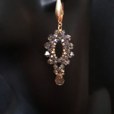 Długie kolczyki wieczorowe wykonane ręcznie z połyskujących kryształków w kolorze dymnego brązu oraz złotych bigli. Model wyróżnia się eleganckim połyskiem i klasycznym kształtem kropli, idealnym do stylizacji na wesele, bal lub imprezę. Biżuteria doda blasku każdemu wyjściu.