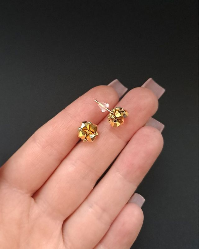 Złote kolczyki sztyfty z czeskich kryształów Preciosa Bicone Crystal Aurum i koralików Miyuki Delica. Drobne kolczyki handmade o średnicy 0,7 cm z fasetowanymi kryształami o metalicznym połysku, złotą bazą ze stali nierdzewnej i przestrzenną formą.