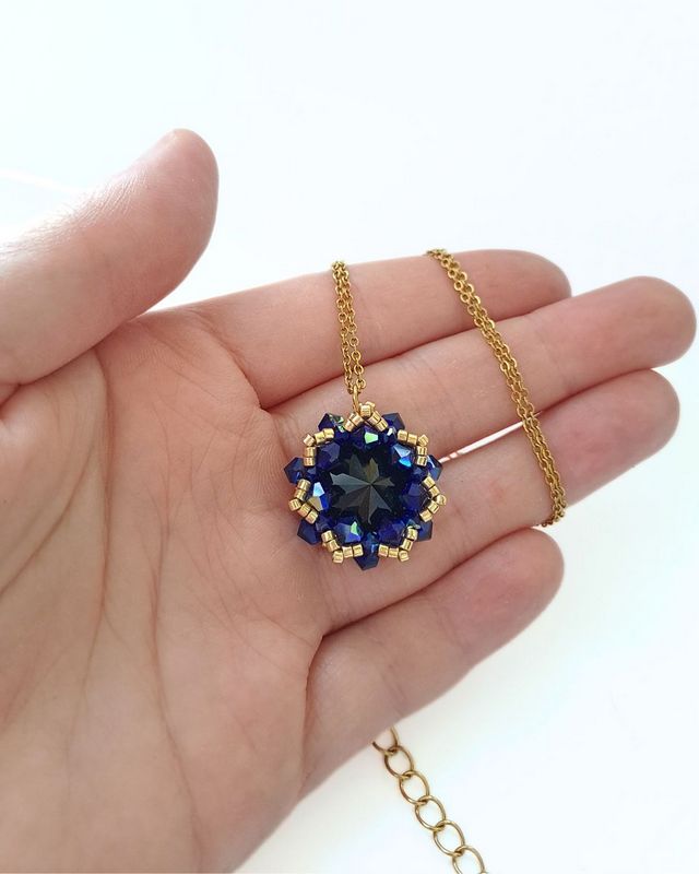 Złoty naszyjnik Cobalt Maxima z czarnym kryształem Preciosa Rivoli Maxima Jet otoczonym kobaltowymi kryształkami Bicone Cobalt AB. Ręcznie wyplatana zawieszka o wymiarach około 2,7 × 2,9 cm ze złotymi koralikami Miyuki Delica na łańcuszku ze stali nierdzewnej.